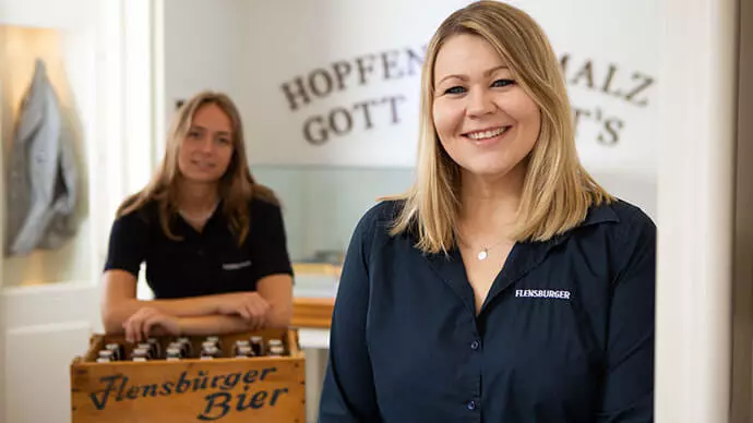 Zwei Mitarbeiterinnen der Flensburger Brauerei in blauen Blusen und mit einer historischen Holzkiste FLENS.