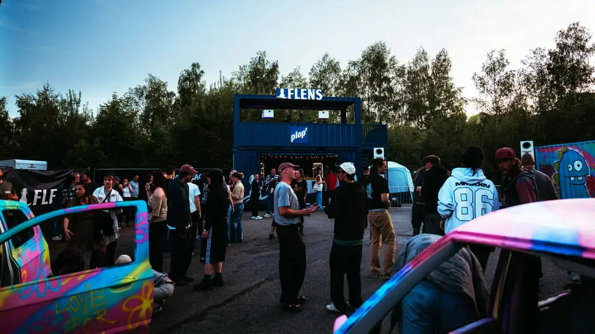 Butcher-Jam-Festival 2026 mit Menschen vor Containerbühne 'FLENS' und bunt besprühten Autos, Abenddämmerung.