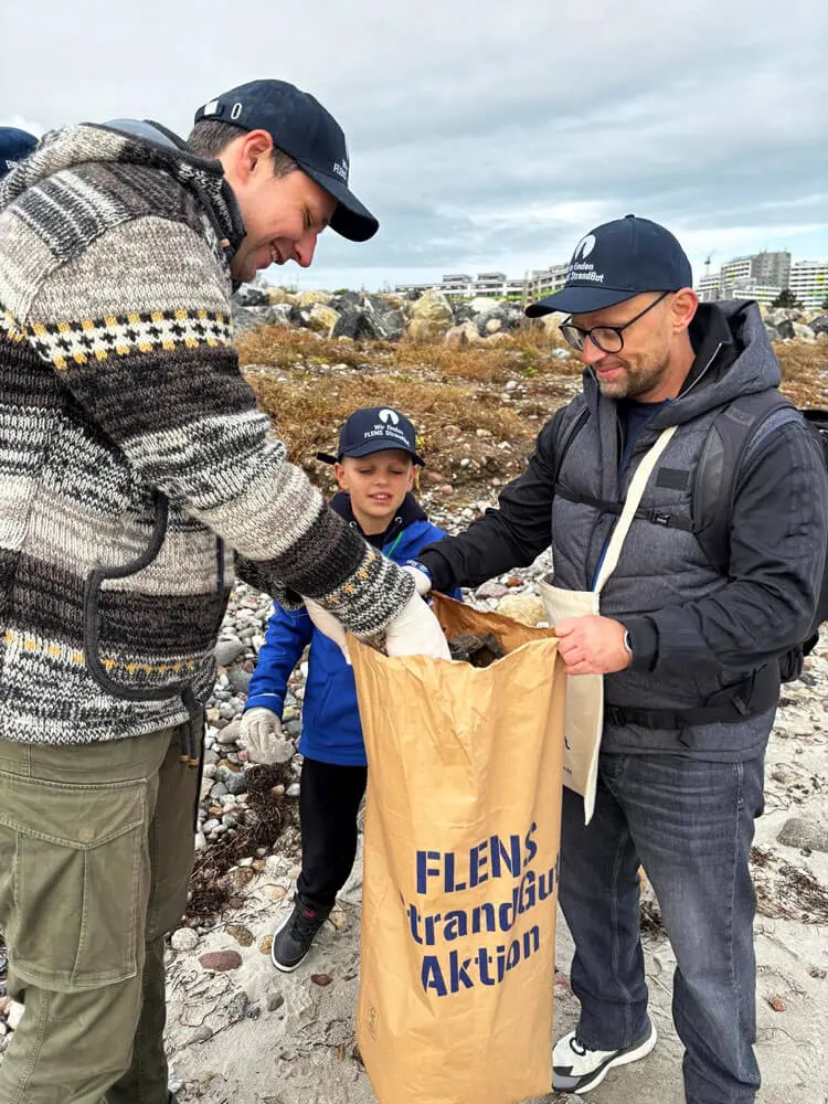 Drei Personen bei einer FLENS Strandreinigungsaktion sammeln Müll in einem Sack.