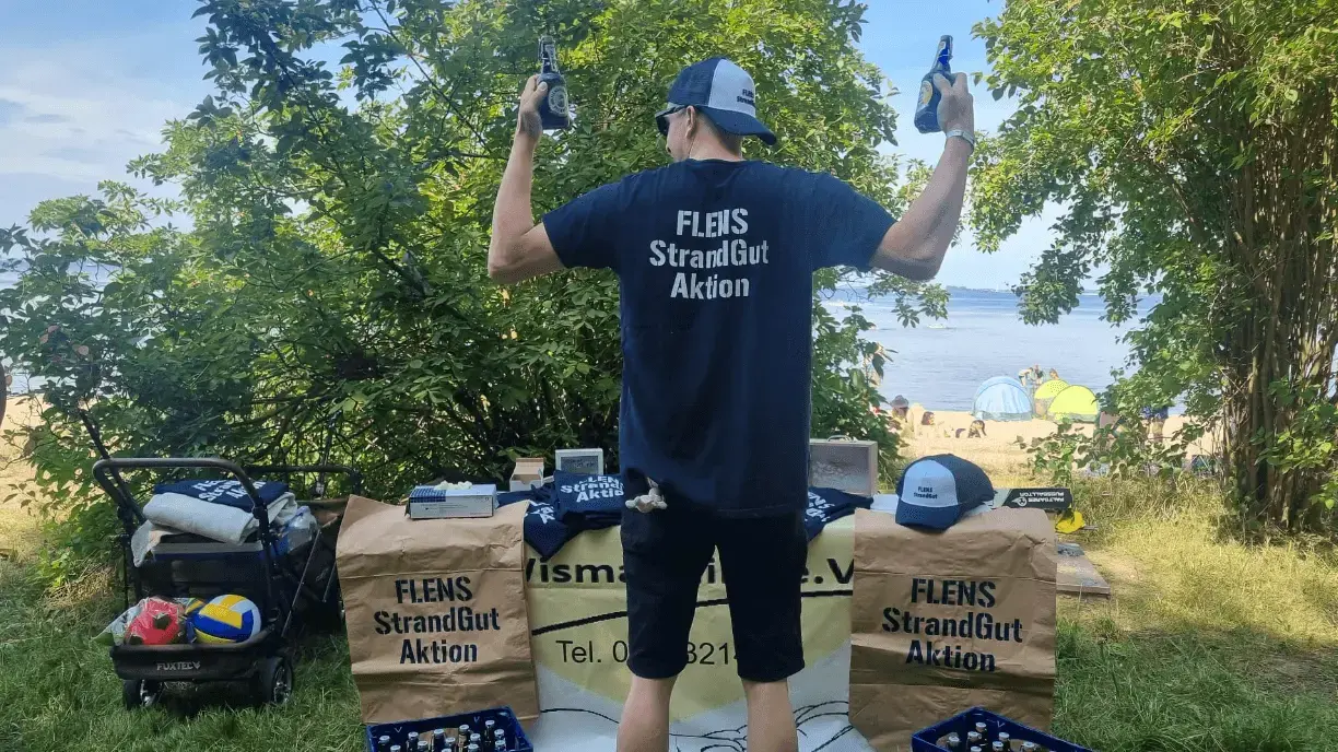 Mann hält Flaschen hoch bei FLENS Strandgut Aktion am sonnigen Strand.