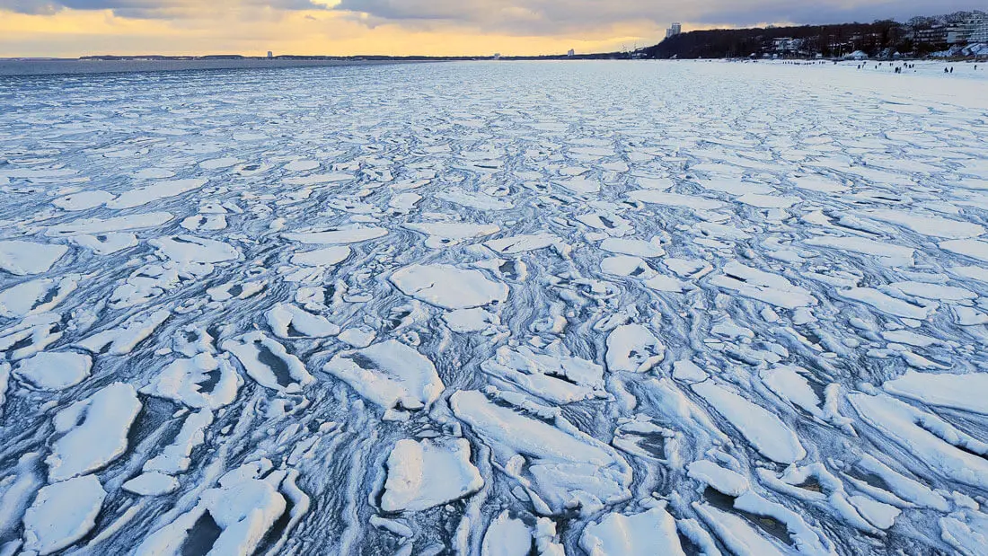 Vereiste Küste mit treibenden Eisschollen und Schneeschichten im Winter bei Sonnenuntergang.