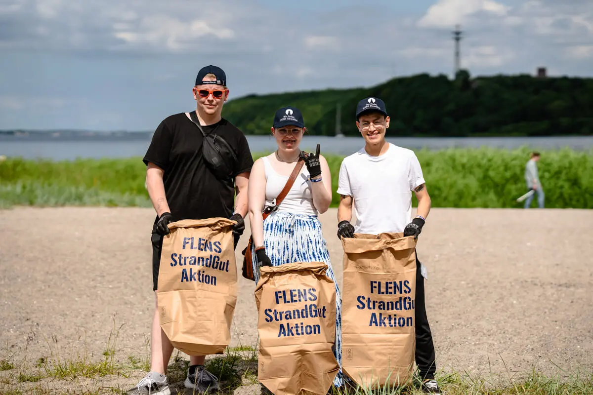 Drei Freiwillige mit Müllsäcken bei einer FLENS Strandreinigungsaktion am Strand in Flensburg.