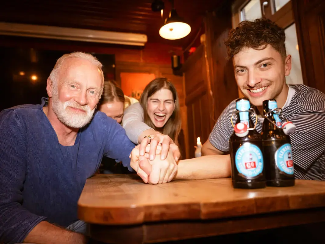 Ein älterer und ein jüngerer Mann und eine Frau stapeln ihre Hände übereinander, im Vordergrund zwei Flaschen Flensburger Strand-Lager 0,0 %.