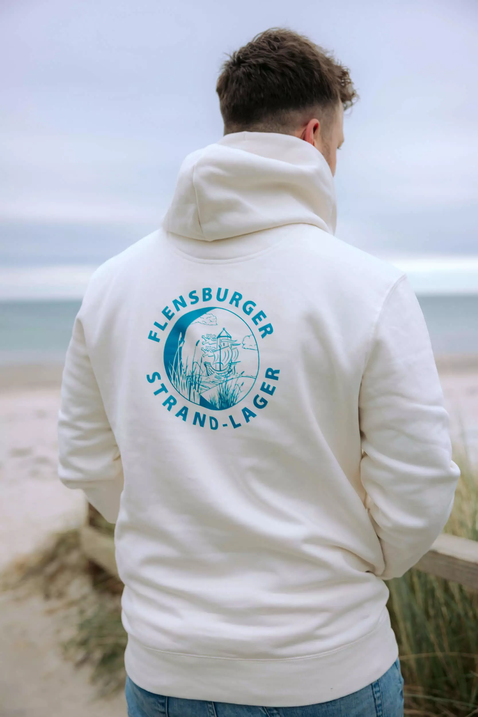 Person am Strand mit Strand-Lager-Hoodie von FLENS: cremeweißer Stoff mit türkisblauem Logo.