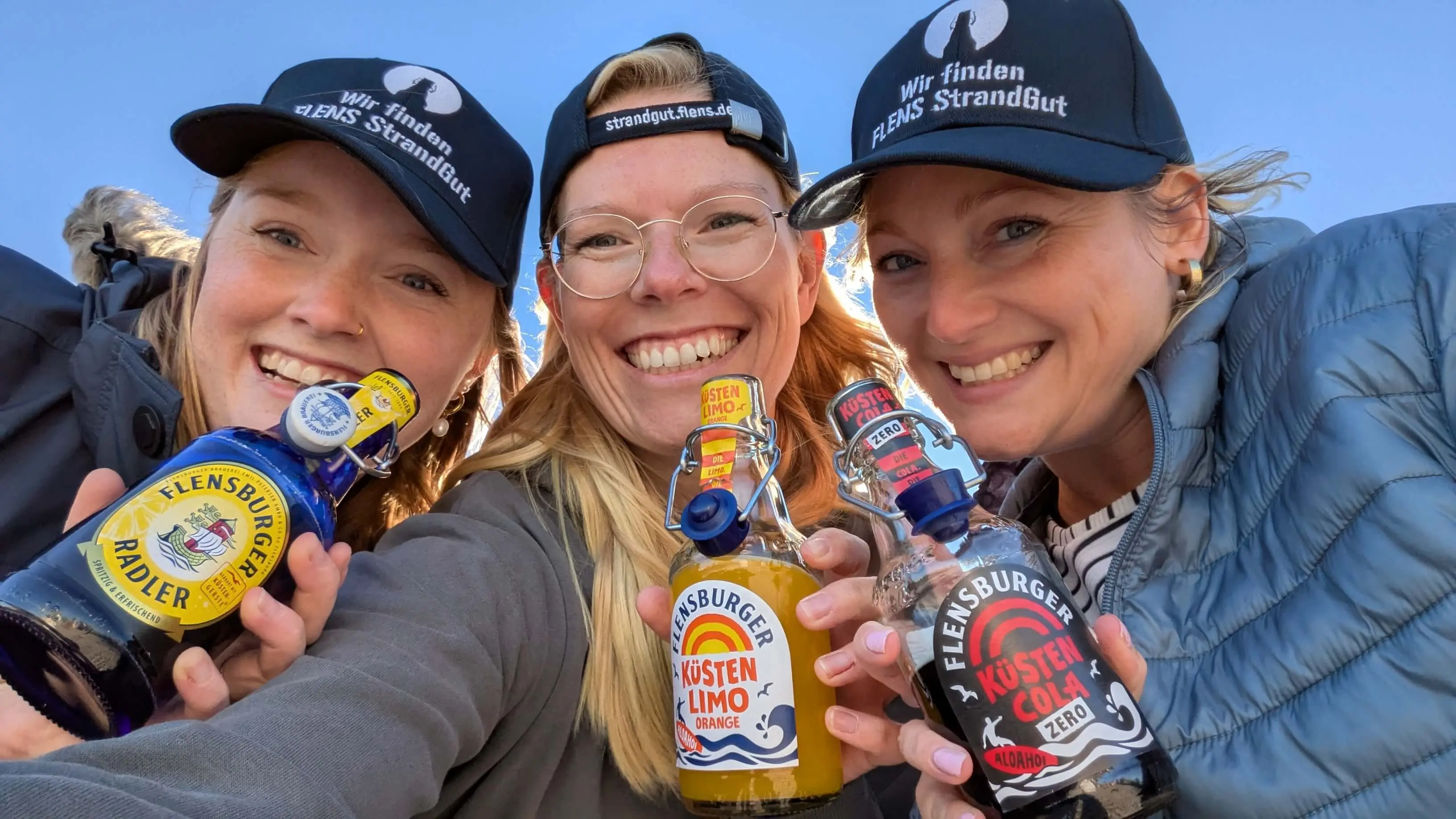 Drei fröhliche Frauen halten Flensburger Getränke in die Kamera, tragen FLENS StrandGut Caps.