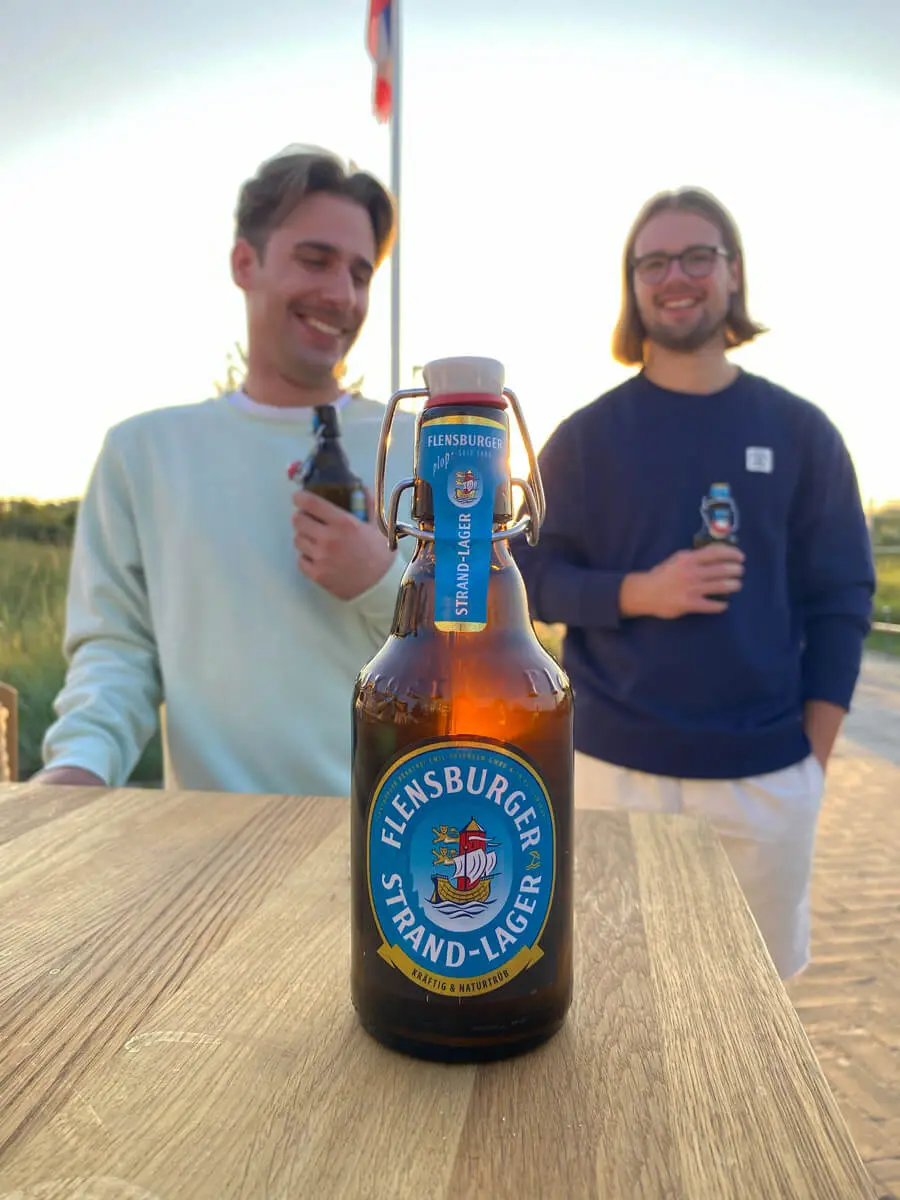Flensburger Strand-Lager Flasche auf Holztisch, zwei Personen mit Bier im Hintergrund am Strand bei Sonnenuntergang.