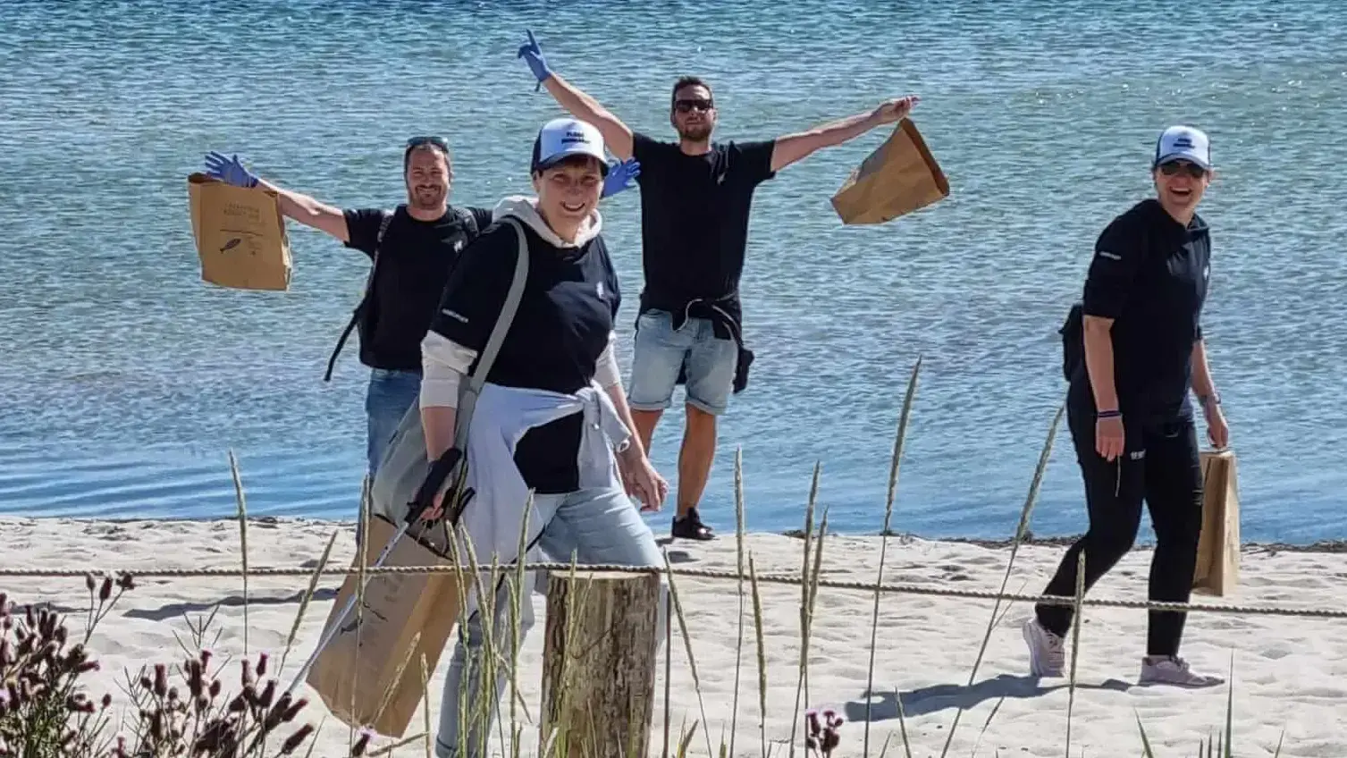 Gruppe von Freiwilligen reinigt Strand mit Müllsäcken vor Hafenkulisse.