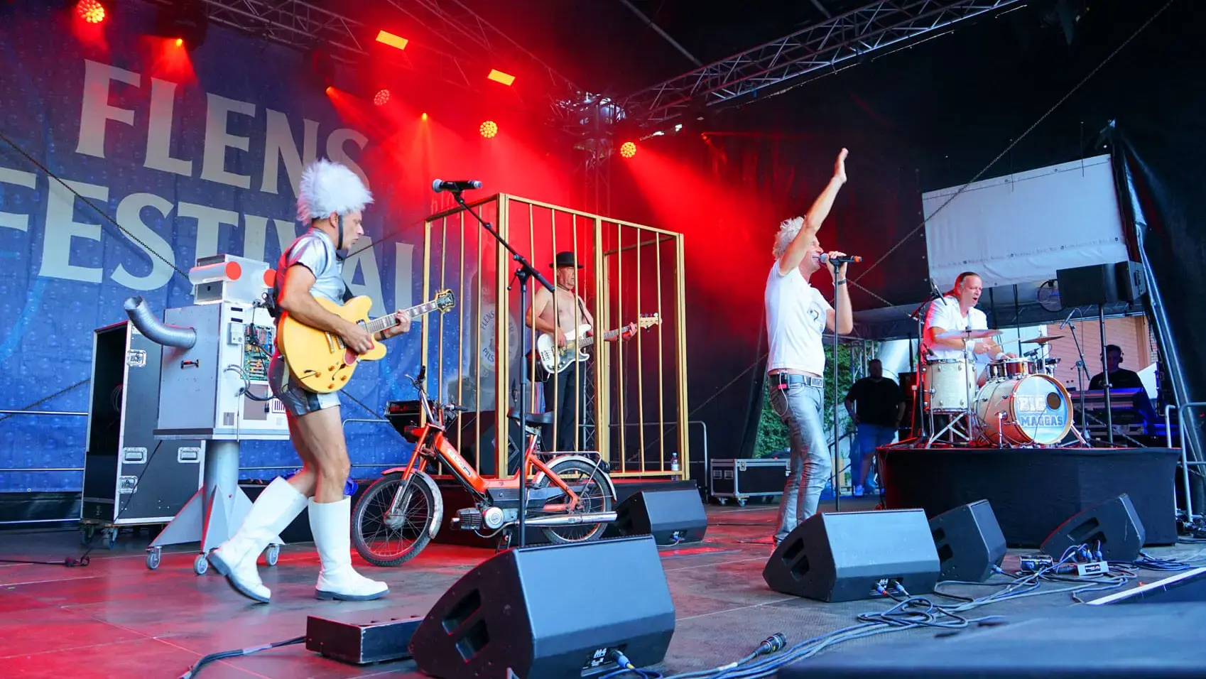 Die BIG MAGGAS Coverband in Aktion auf der Bühne vom FLENS Festival. Links der Gitarrist im Silberhemd und weißer Flausch-Mütze, dann der Bassist im Goldkäfig und Melone, dann der Frontsänger winkend und im weißen Shirt und rechts der Schlagzeuger am Drumset. Außerdem ein selbstgebauter Roboter und ein Moped auf der Bühne.