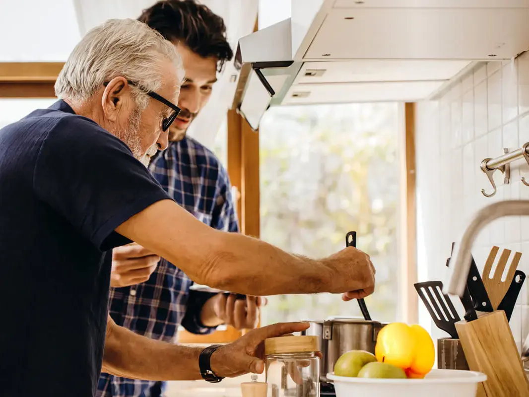 Senior und Erwachsener kochen gemeinsam in heller Küche, rühren Topf neben Gemüseschale und Kochutensilien.