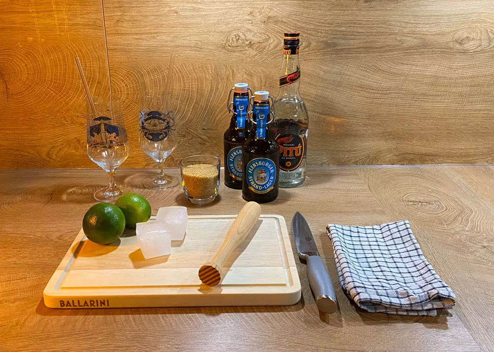 Übersicht über die Utensilien, die man für den Biercocktail Caibierinha benötigt: Zwei Gäser, braunen Zucker, Flensburger Strand-Lager, weißen Rum, Limetten, einen Stößel, Eis, ein Messer, ein Holzbrett und optional ein Küchenhandtuch.