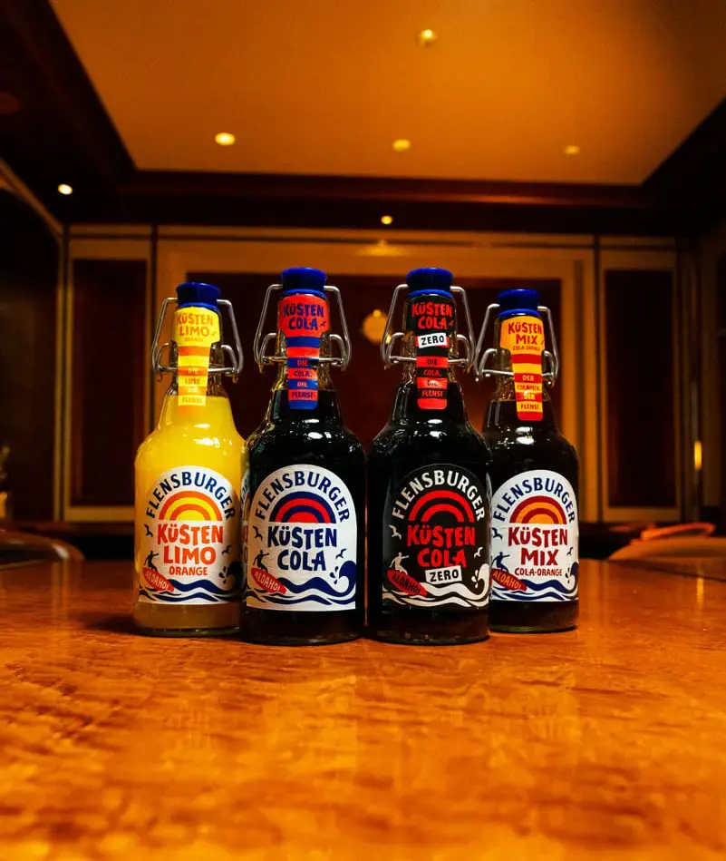 Vier Flensburger Küstenlimo-Flaschen (Küstenlimo Orange, Küstencola, Küstencola Zero, Küstenmix) auf Holztisch.