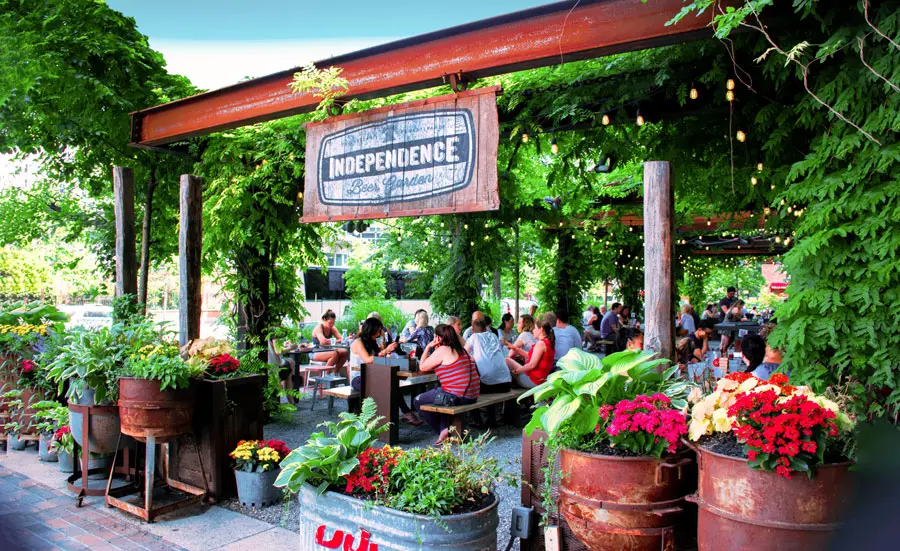 Belebter Biergarten mit grüner Pergola, Lichterketten, Blumen und Gästen; Schild mit "Beer Garden"-Aufschrift hängt über dem Eingang.