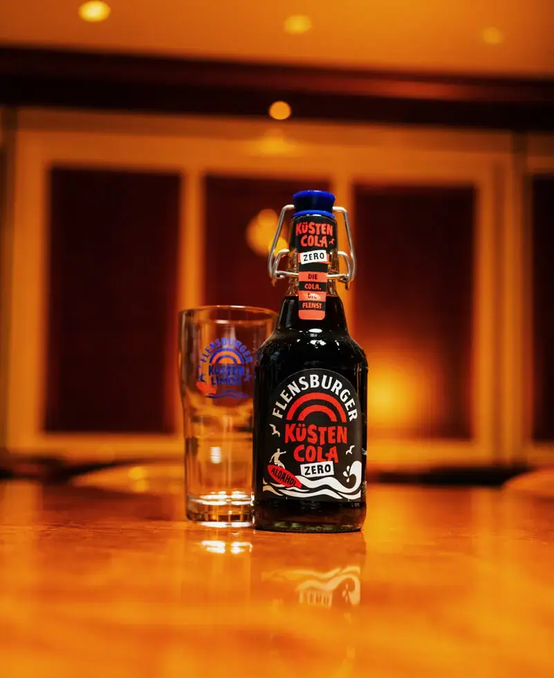 Flensburger-Küstencola-Zero-Flasche mit Küstenlimo-Glas auf Holztheke im warmen Barlicht.