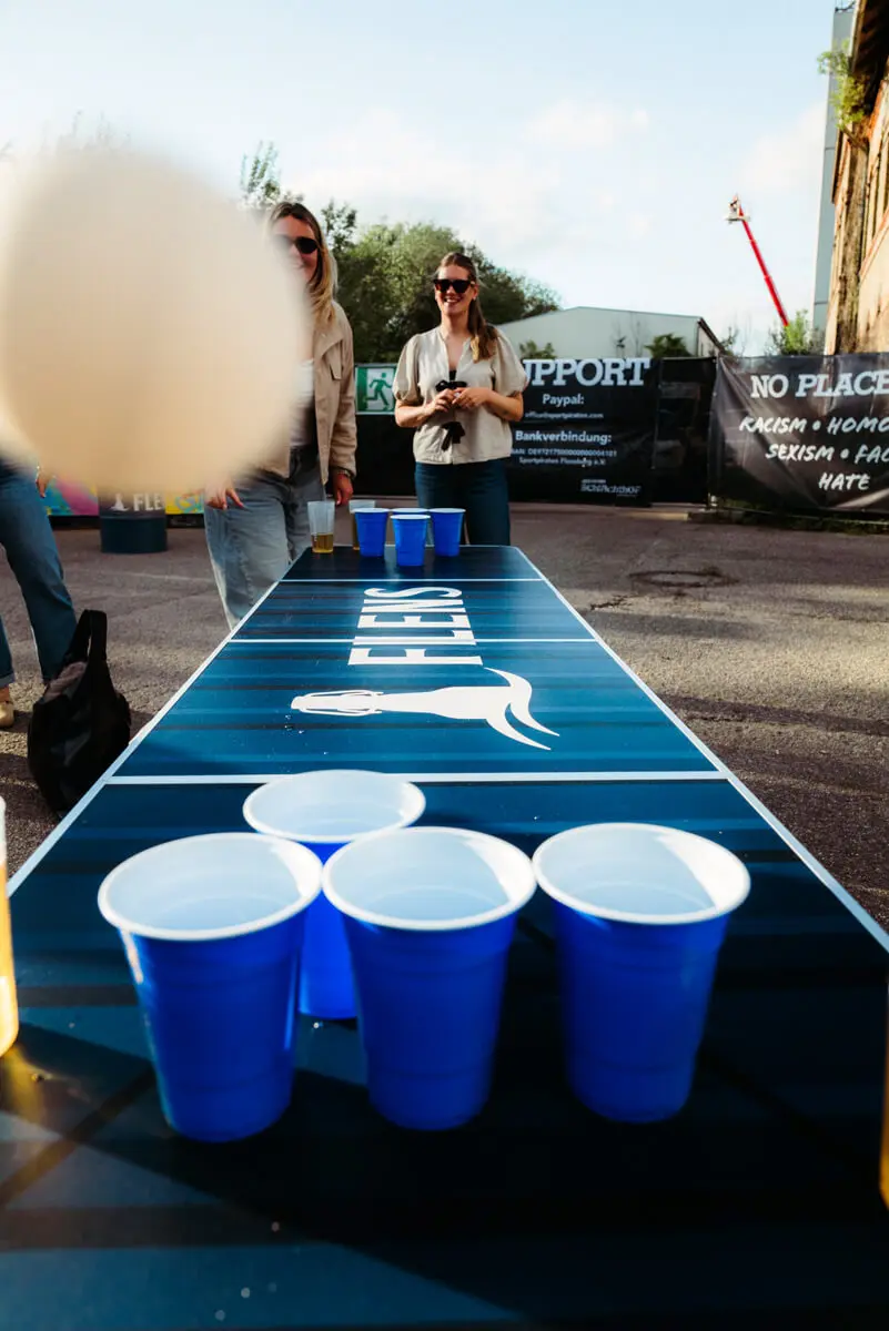 Outdoor-Beerpong: blaue Plastikbecher auf Tisch, Spieler und Zuschauer im Hintergrund.