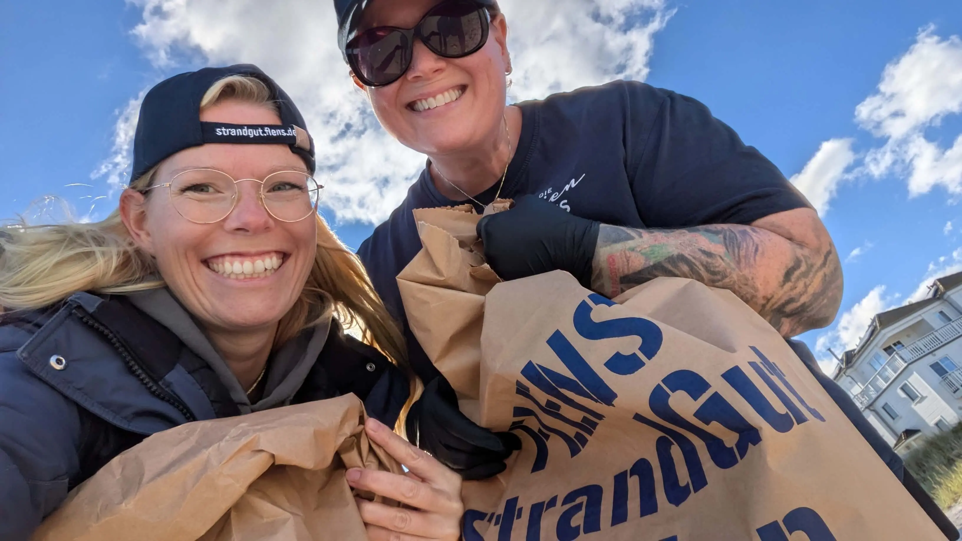 Zwei lächelnde Frauen bei FLENS Müllsammelaktion am Strand mit Müllsäcken in den Händen.
