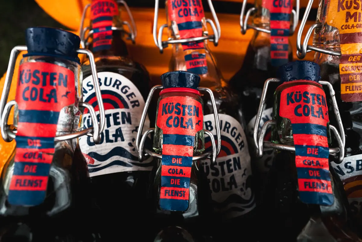 Küstencola-Flaschen mit Bügelverschluss und roten-blauen Etiketten, Nahaufnahme einer Kühlbox.