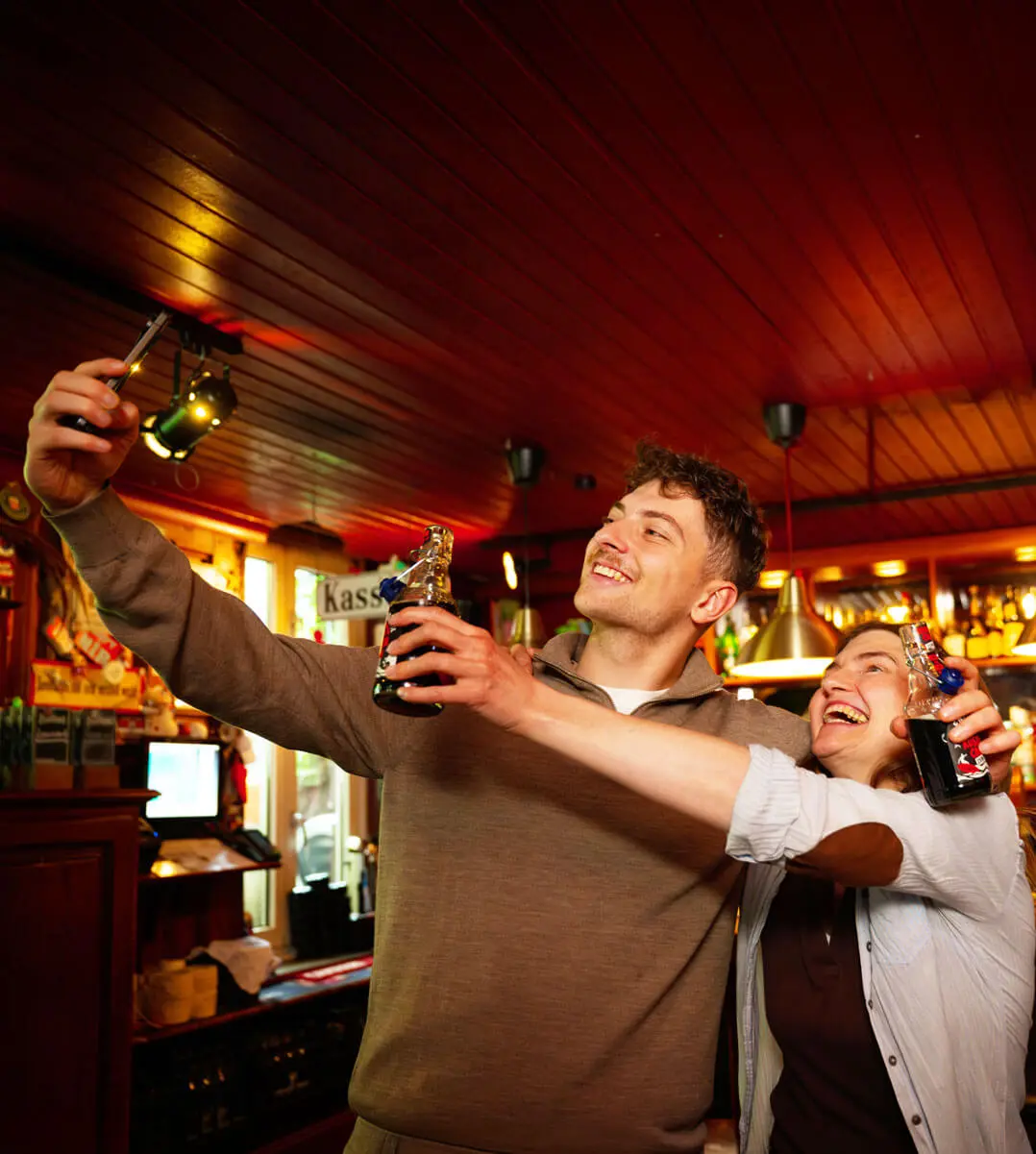 Zwei Freunde machen ein Selfie an der Bar, lachen und halten Bügelverschlussflaschen mit Küstencola Zero in gemütlicher, warm beleuchteter Kneipenatmosphäre.