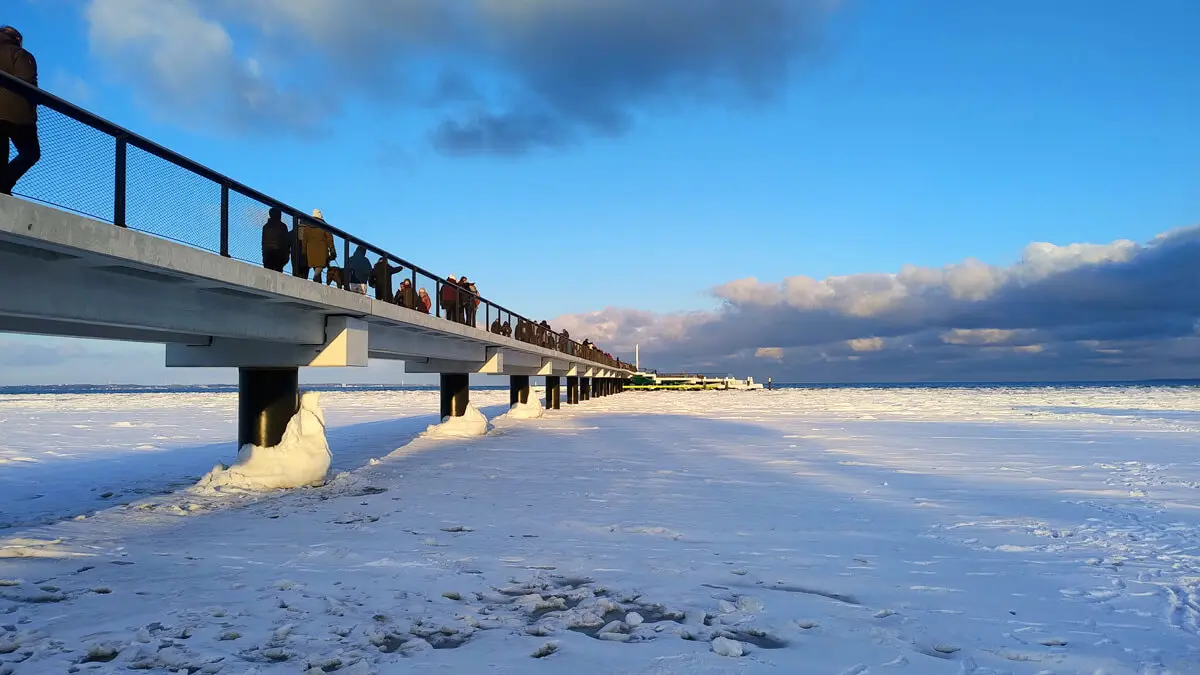 Seebrücke mit Spaziergängern über zugefrorenem, verschneitem Strand unter blauem Winterhimmel.