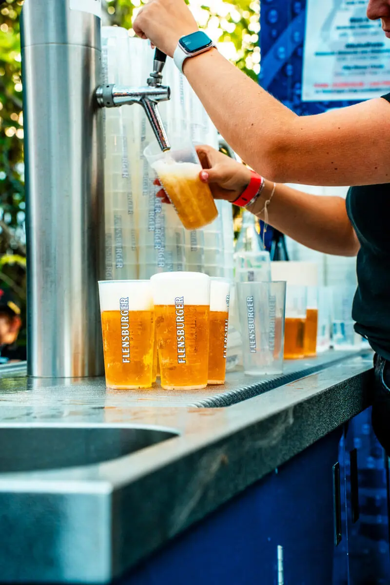 Person zapft Flensburger Bier aus Zapfhahn in Plastikbecher mit appetitlichem Schaum.