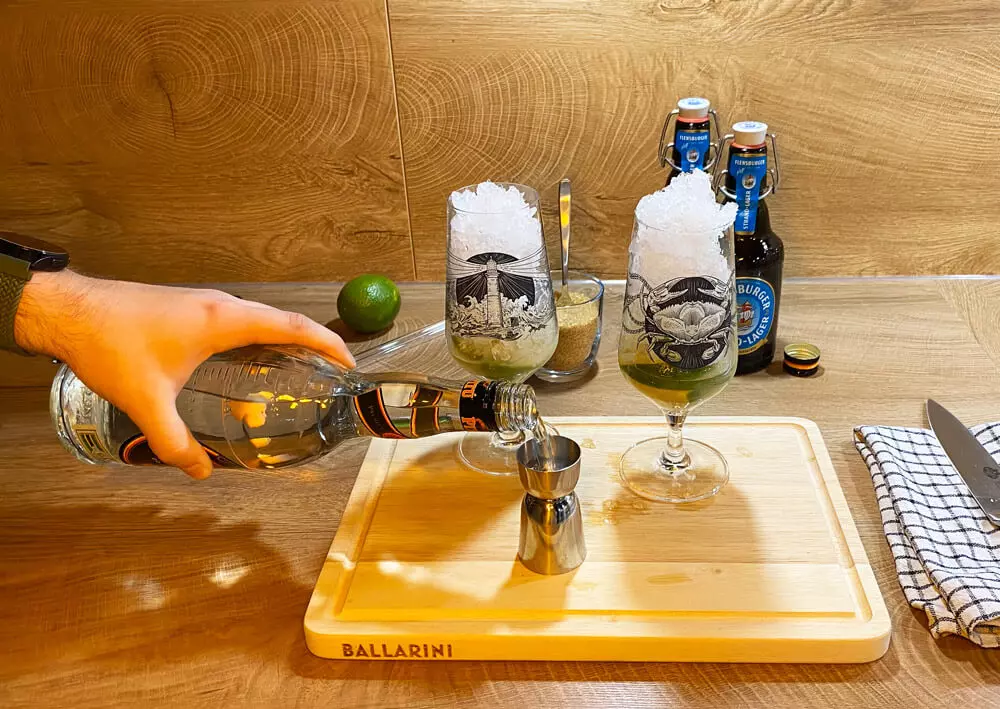 Weißer Rum wird in einen Messbecher gefüllt, dahinter Cocktailutensilien.