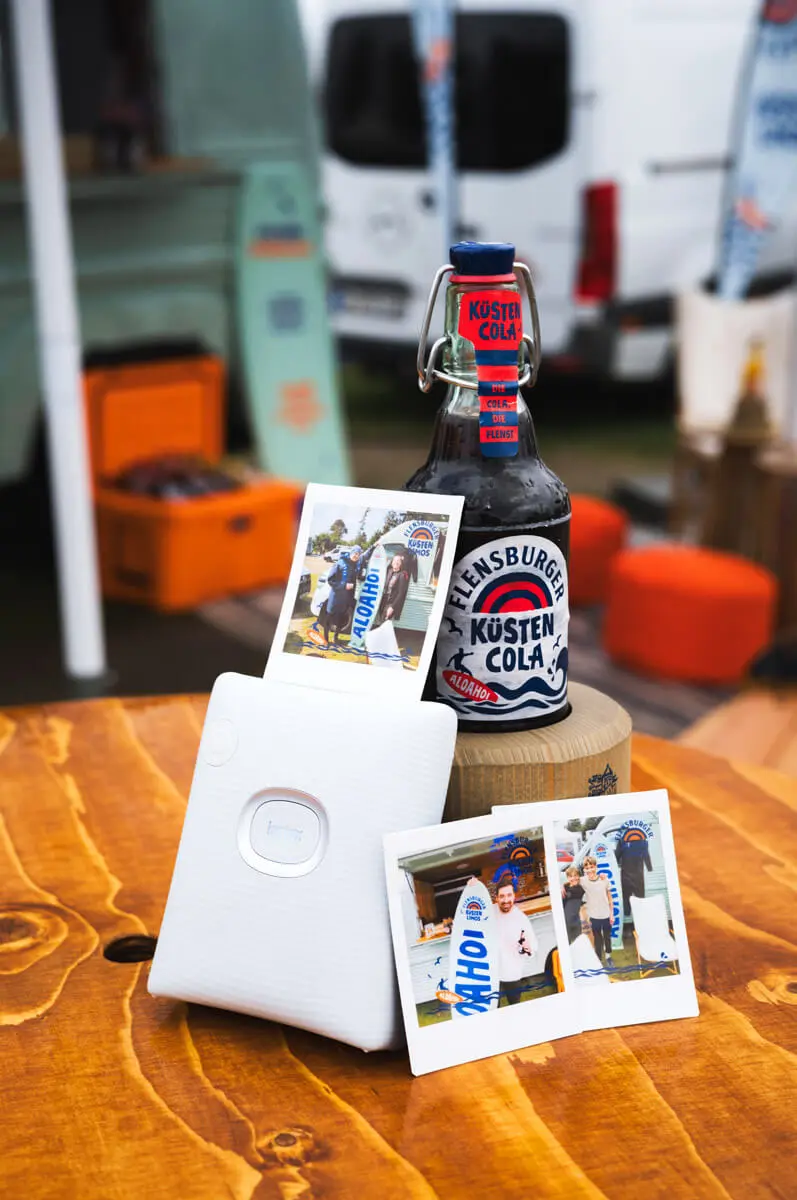 Flensburger Küstencola-Flasche mit Polaroids und Mini-Fotodrucker auf Holztisch.