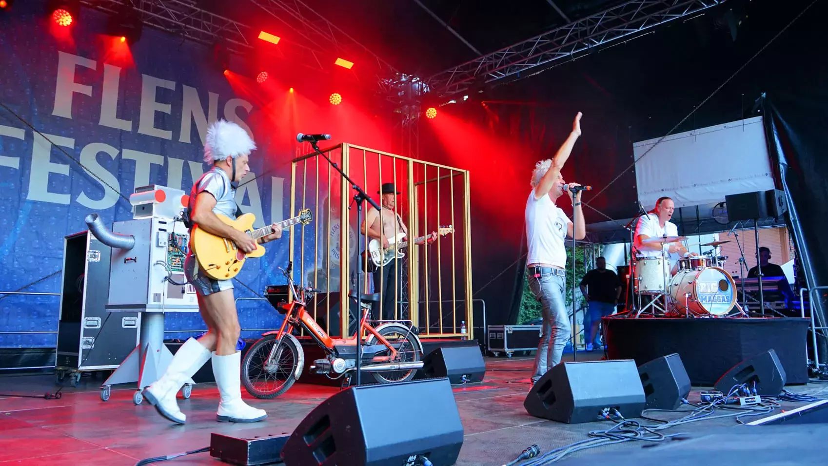 Die BIG MAGGAS Coverband in Aktion auf der Bühne vom FLENS Festival. Links der Gitarrist im Silberhemd und weißer Flausch-Mütze, dann der Bassist im Goldkäfig und Melone, dann der Frontsänger winkend und im weißen Shirt und rechts der Schlagzeuger am Drumset. Außerdem ein selbstgebauter Roboter und ein Moped auf der Bühne.