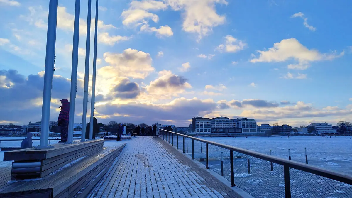 Verschneiter Holzsteg an gefrorener Ostsee unter blauem Winterhimmel, Menschen auf Promenade vor Ufergebäuden.