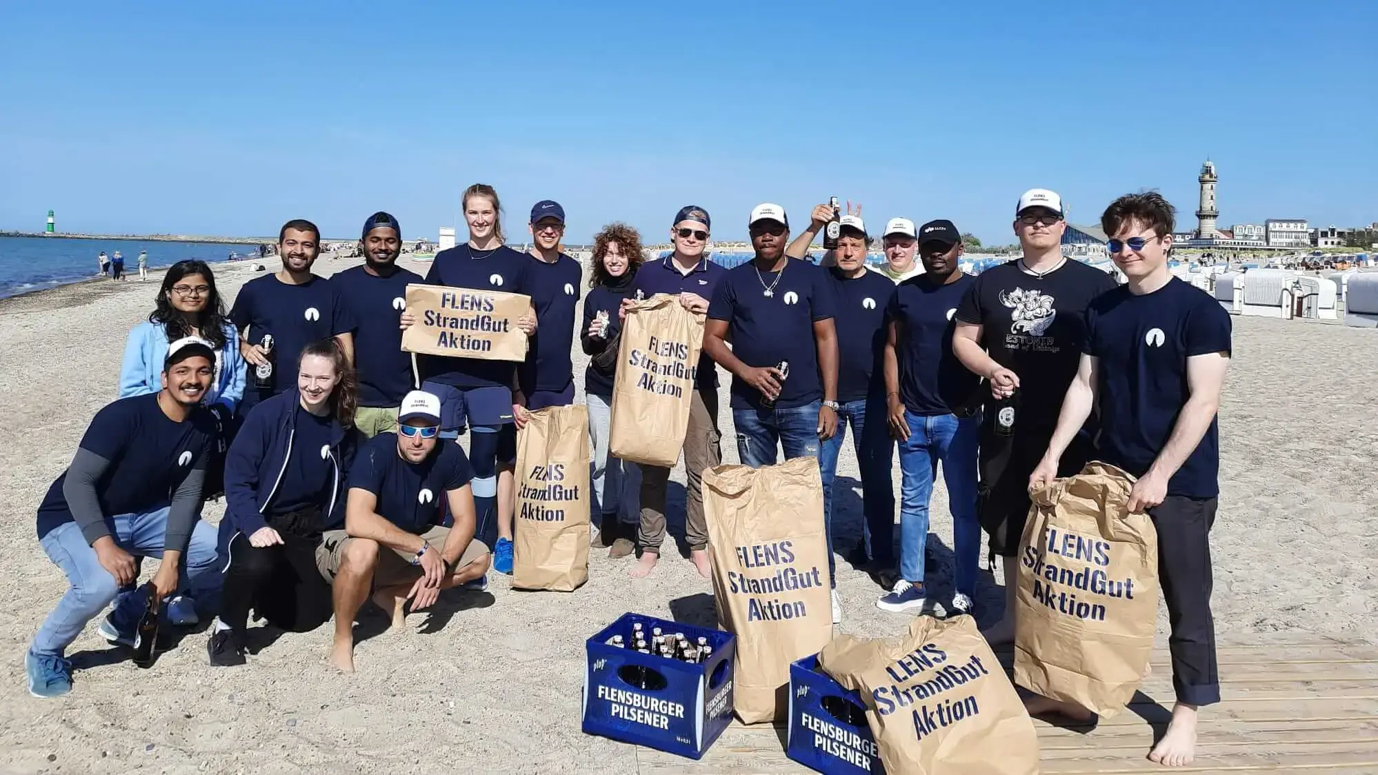 Gruppe von Freiwilligen bei einer FLENS Strandreinigungsaktion mit Säcken am sonnigem Strand.