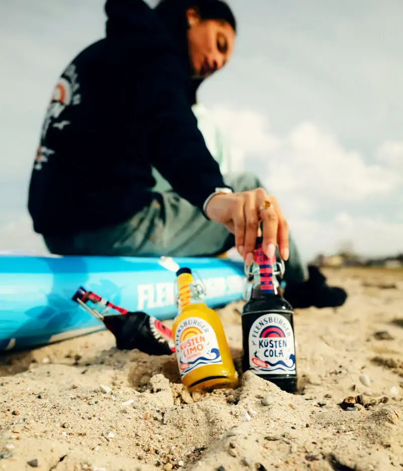 Strandszene: Flensburger Küstencola und Küstenlimo im Sand, Person auf blauem SUP greift nach der Cola.