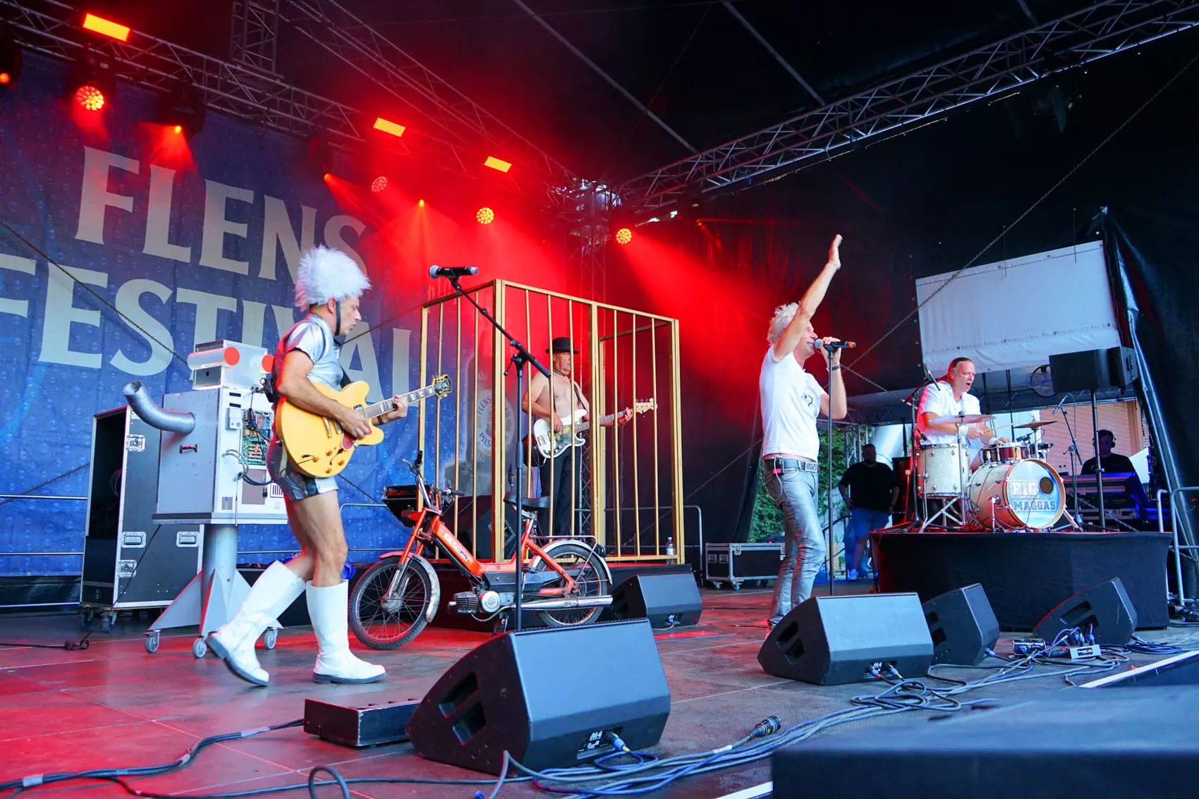 Die BIG MAGGAS Coverband in Aktion auf der Bühne vom FLENS Festival. Links der Gitarrist im Silberhemd und weißer Flausch-Mütze, dann der Bassist im Goldkäfig und Melone, dann der Frontsänger winkend und im weißen Shirt und rechts der Schlagzeuger am Drumset. Außerdem ein selbstgebauter Roboter und ein Moped auf der Bühne.