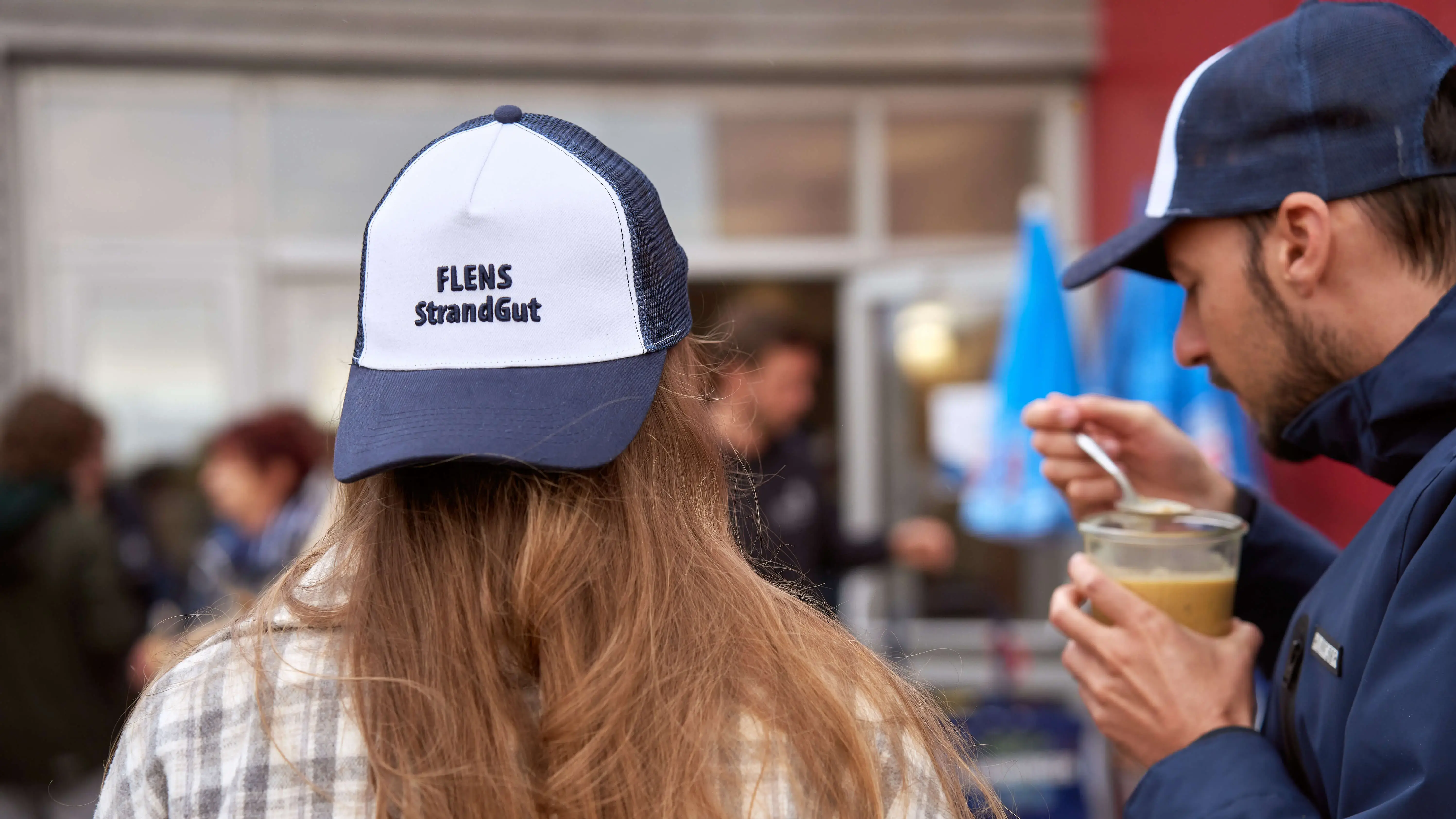 Person trägt Kappe mit Logo "FLENS StrandGut" auf einem belebten Platz.