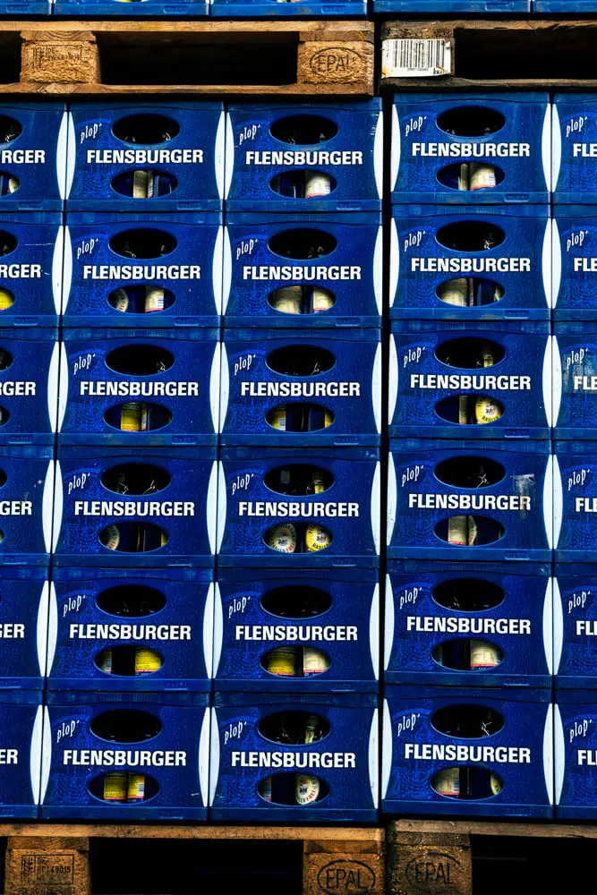 Gestapelte blaue Flensburger Bierkisten auf Paletten mit sichtbaren Flaschen.