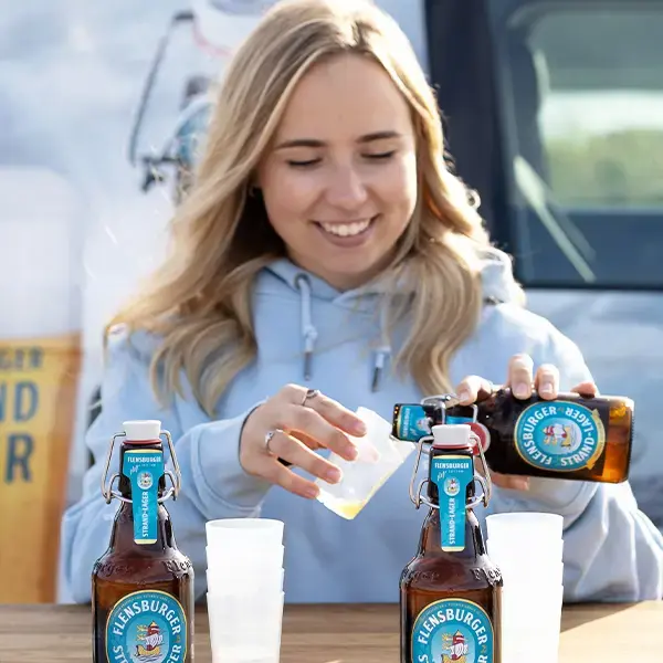 Person schenkt Flensburger-Strand-Lager-Bier in Plastikbecher bei Outdoor-Verkostung vor Promo-Bus.