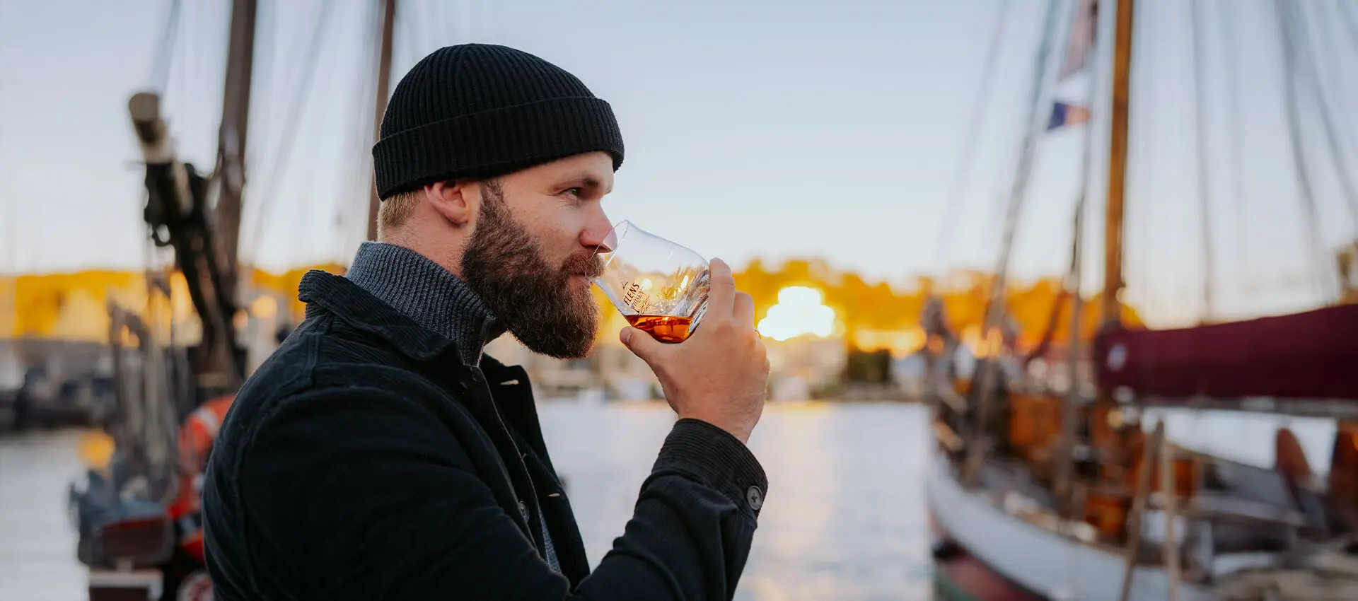 Ein bärtiger Mann trinkt genüsslich einen Schluck FLENS Whisky aus einem Tumbler-Glas im Sonnenuntergang am Hafen von Flensburg.