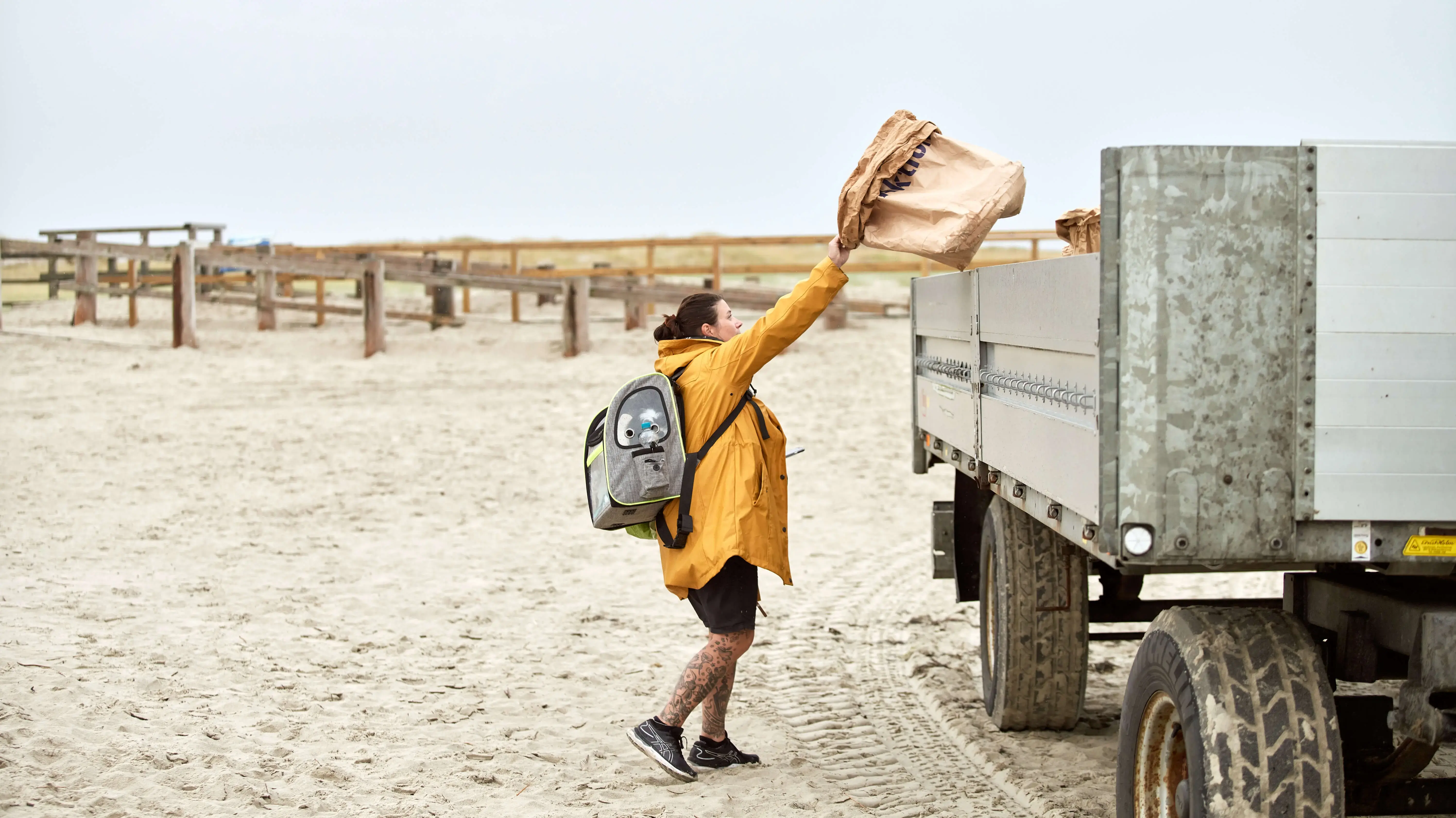Person in gelber Jacke lädt Müllsack auf LKW an einem Strand.