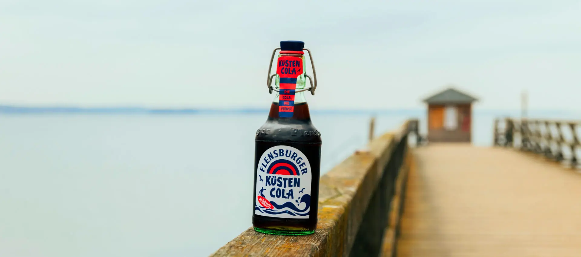 Flensburger Küstencola-Glasflasche auf Holzsteg mit Meer im Hintergrund.