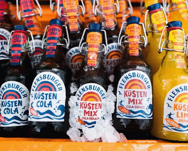 Flensburger Küstenmix auf Eis im Kühlkasten, daneben Flensburger Küstencola und Flensburger Limo Orange.