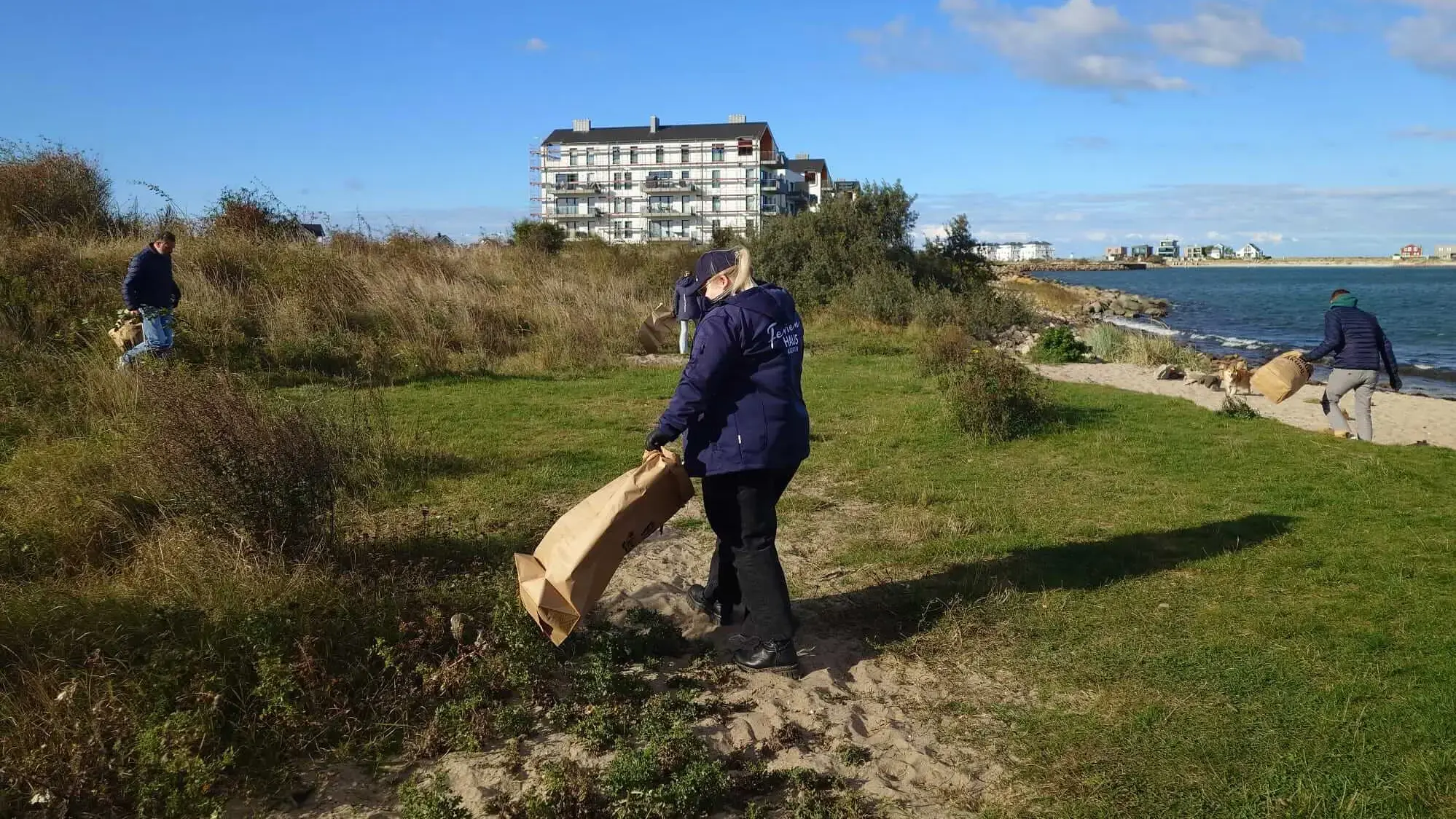 Freiwillige reinigen Strand mit Müllsäcken vor Meeresküste und Gebäuden bei einer FLENS Strandreinigungsaktion.