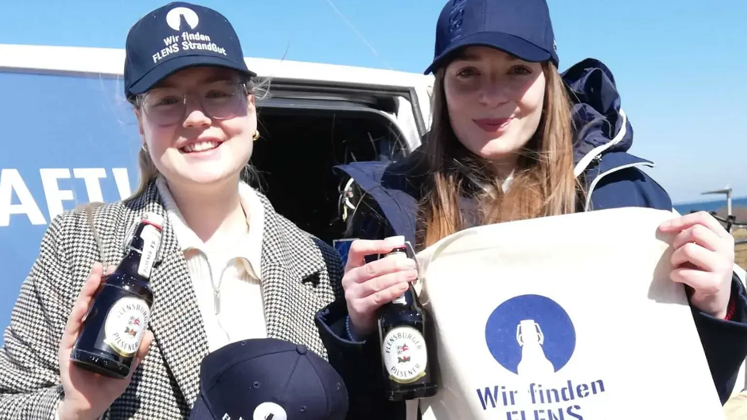 Zwei junge Frauen werben für die FLENS Strandreinigungsaktion am Strand Caps und Jutebeuteln.