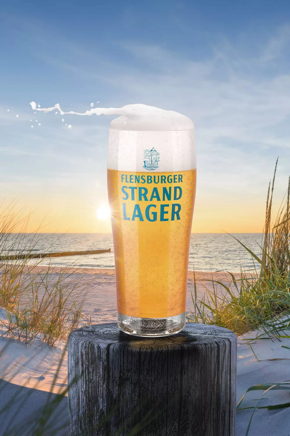 Strand-Lager-Becher mit Flensburger Strand-Lager-Bier gefüllt auf einem Holzpoller vor einer Strandlandschaft.