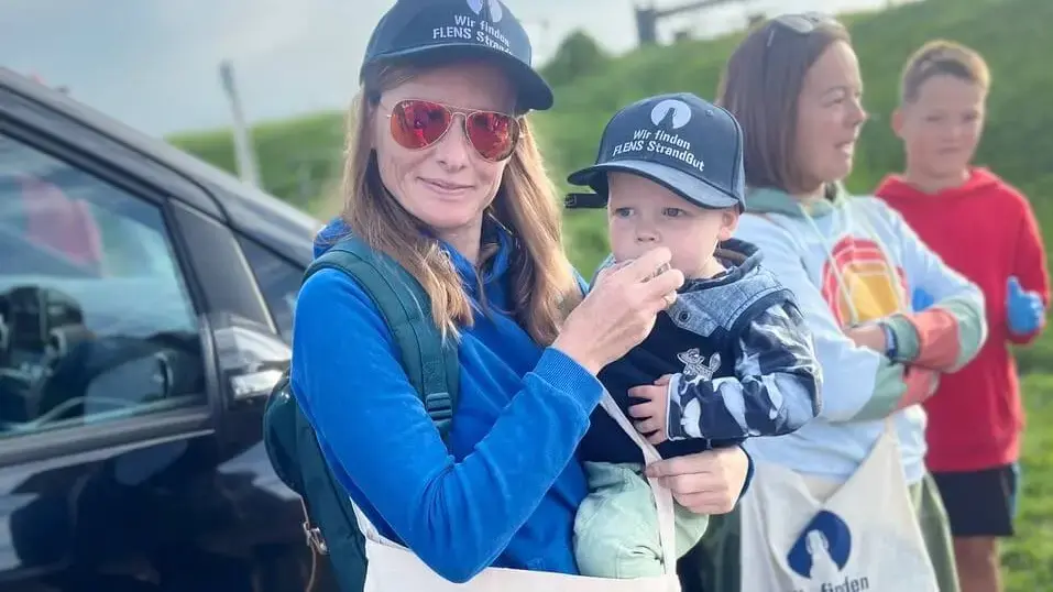Frau und Kind mit "Wir finden FLENS Strandgut"-Caps und -Jutebeuteln, weiter Menschen und ein Auto im Hintergrund.