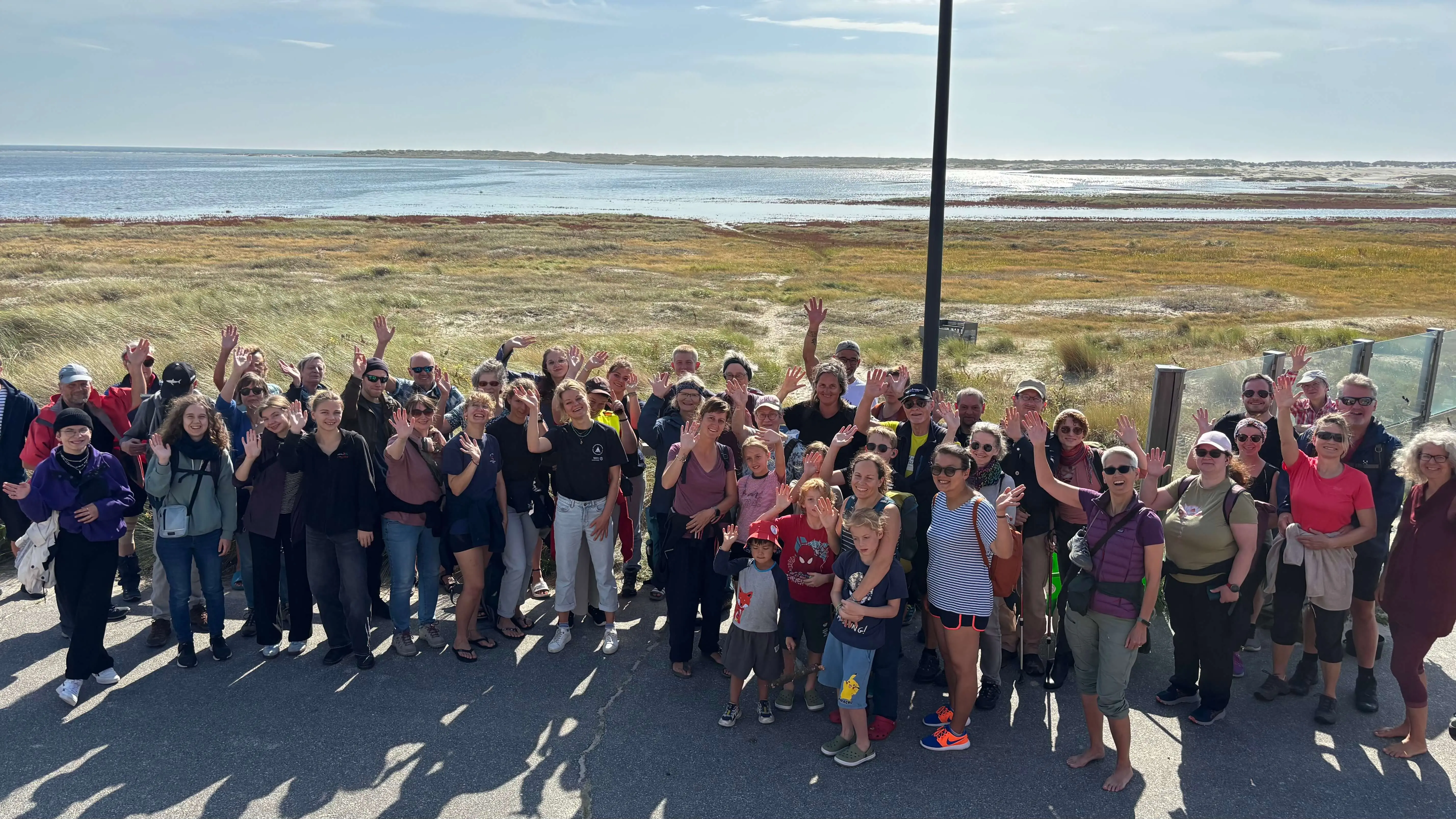 Fröhliche Gruppe von Freiwilligen posiert am sonnigen Küstenstreifen mit ausgestreckten Händen bei einer FLENS StrandGut Aktion.