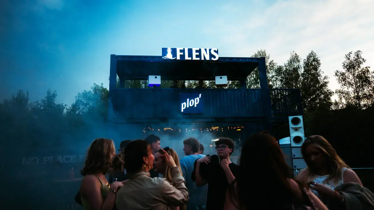 Butcher Jam 2026: Menschen vor Container-DJ-Stage mit Leuchtschrift 'FLENS' und 'plop' bei Abenddämmerung.