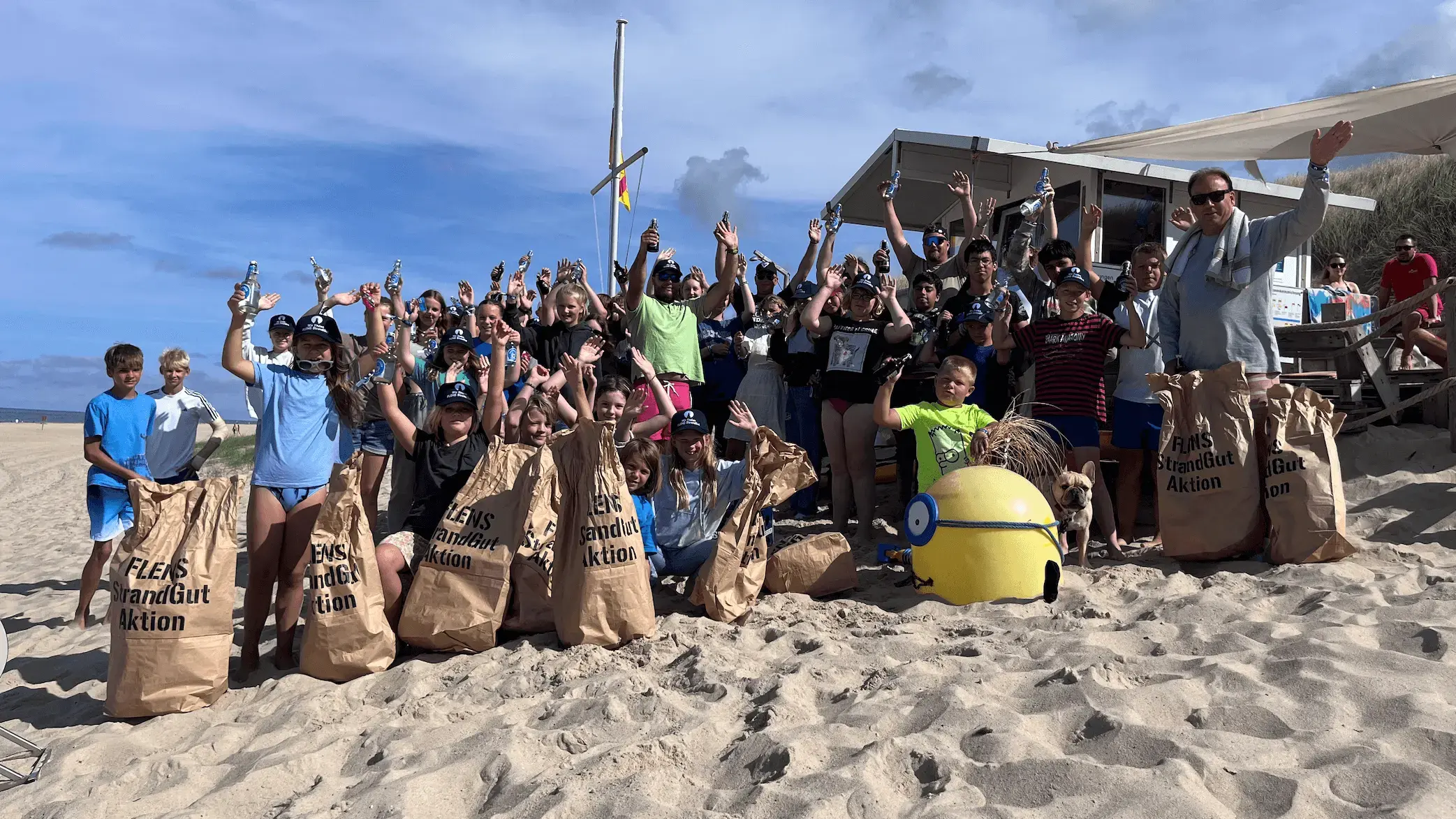Gruppe von Menschen bei einer FLENS Strandreinigungsaktion, jubelnd mit Müllsäcken an einem sonnigen Strand.