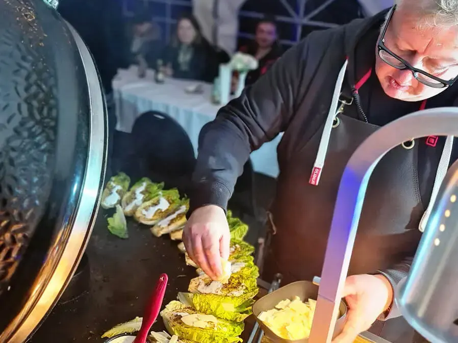 Grillweltmeister Marco Greulich bereitet gegrillten Salat bei der Grillweltmeisterparty von FLENS zu, Gäste im Hintergrund.