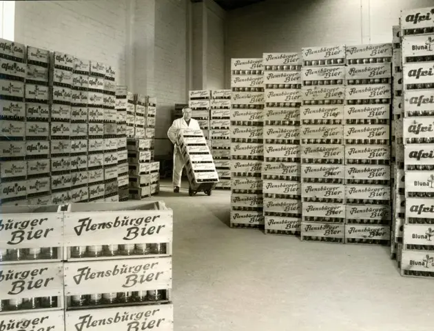 Expedition damals bei FLENS mit gestapelten Flensburger Bierkisten aus Holz und Arbeiter mit einer Sackkarre.