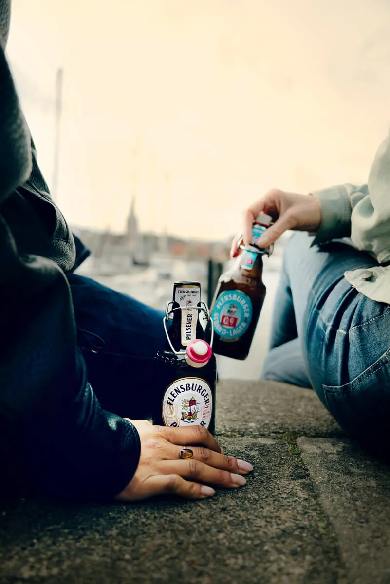 Zwei Personen trinken Flensburger Pilsener am Hafen auf Kaimauer, Bügelflaschen im Vordergrund