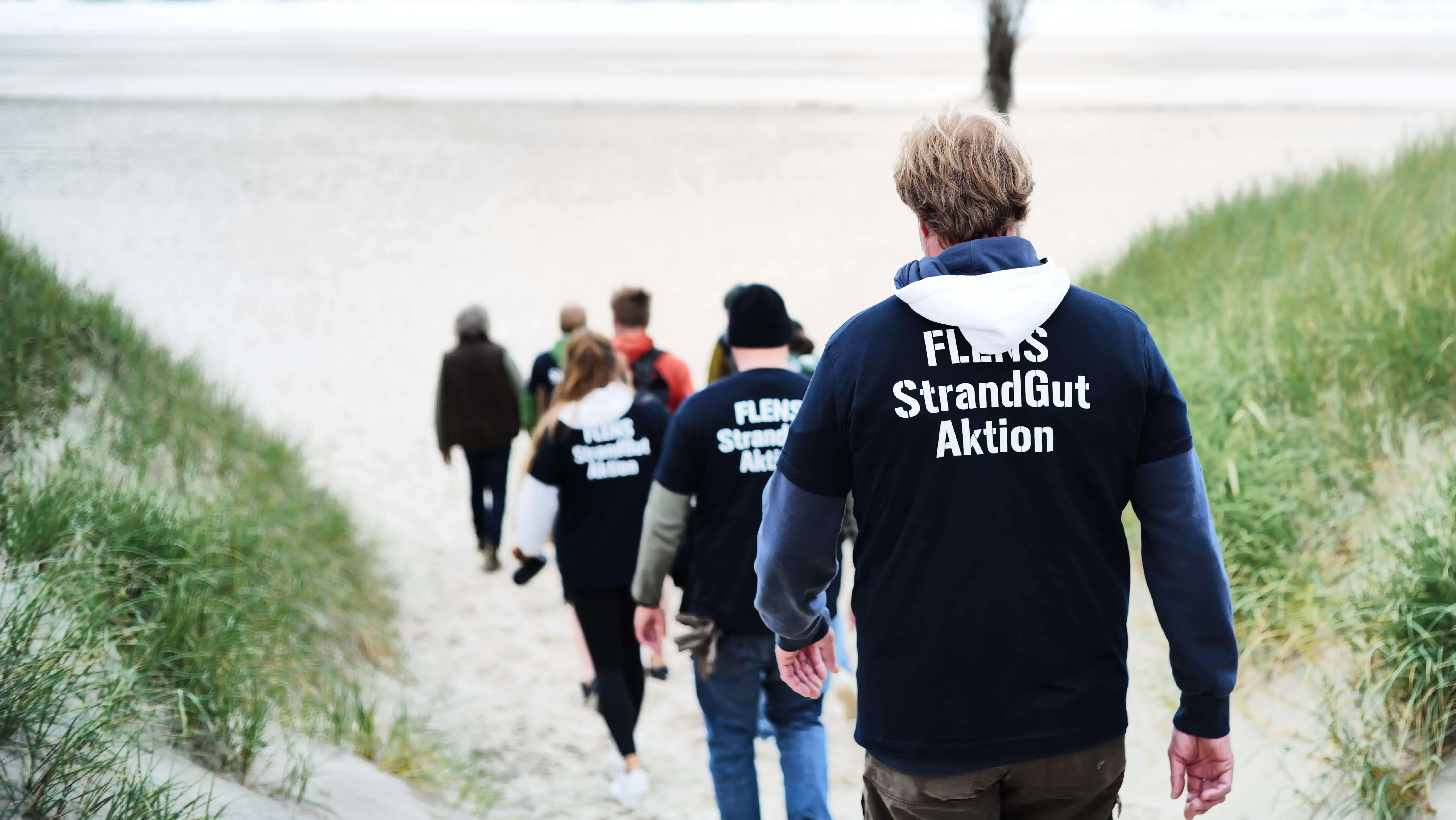 Gruppe in "FLENS StrandGut Aktion" Jacken geht am Strand entlang.
