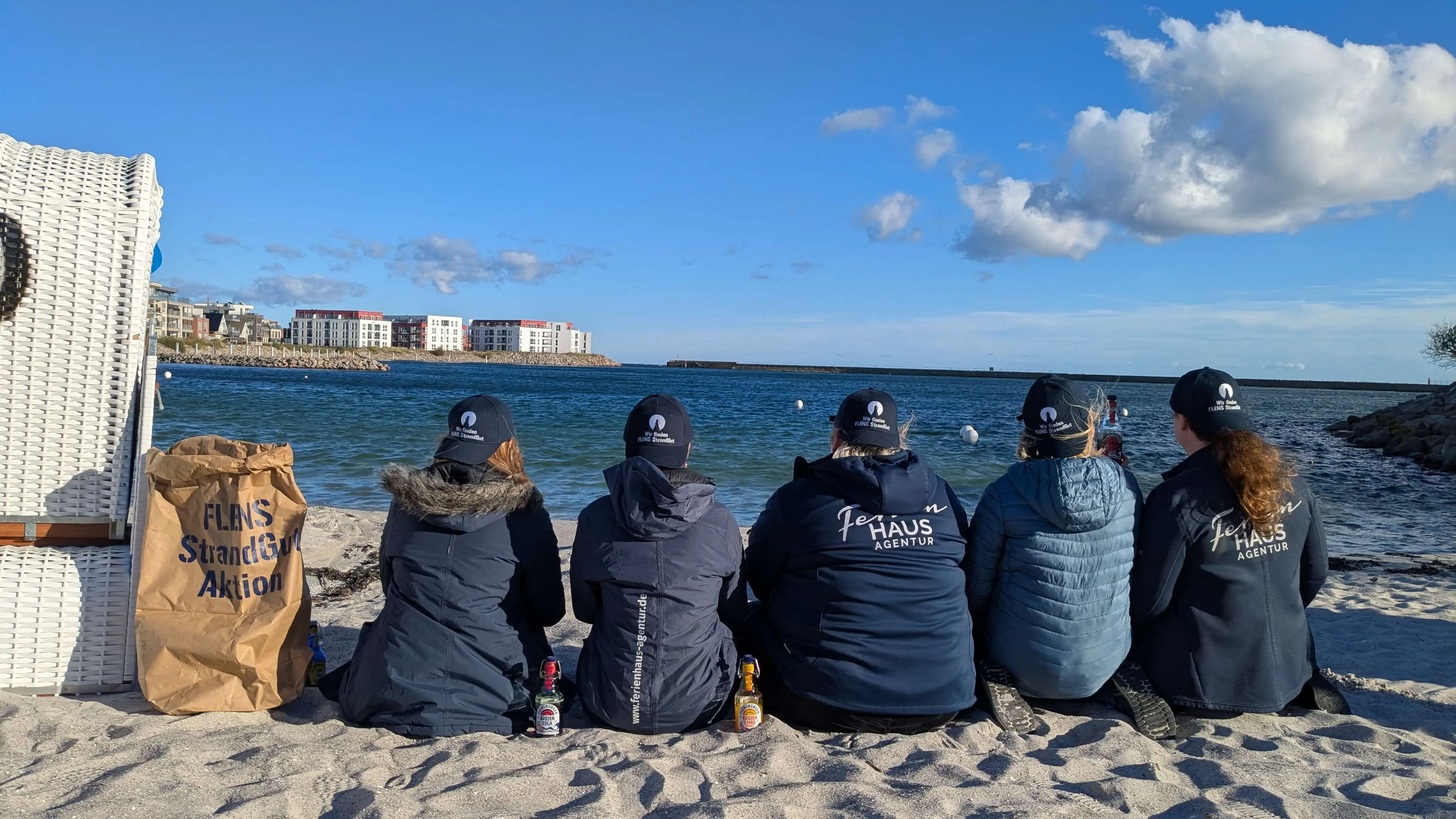 Gruppe von Freiwilligen der FLENS StrandGut Aktion, alle in blau gekleidet, sitzt am Strand und schaut auf das Meer.
