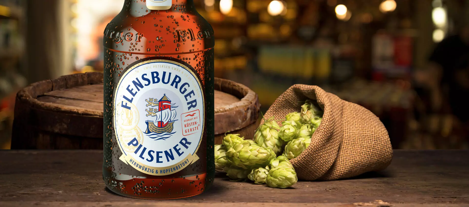 Hopfendolden in einem Jutesack auf einem Tisch, daneben eine Flasche Flensburger Pilsener.